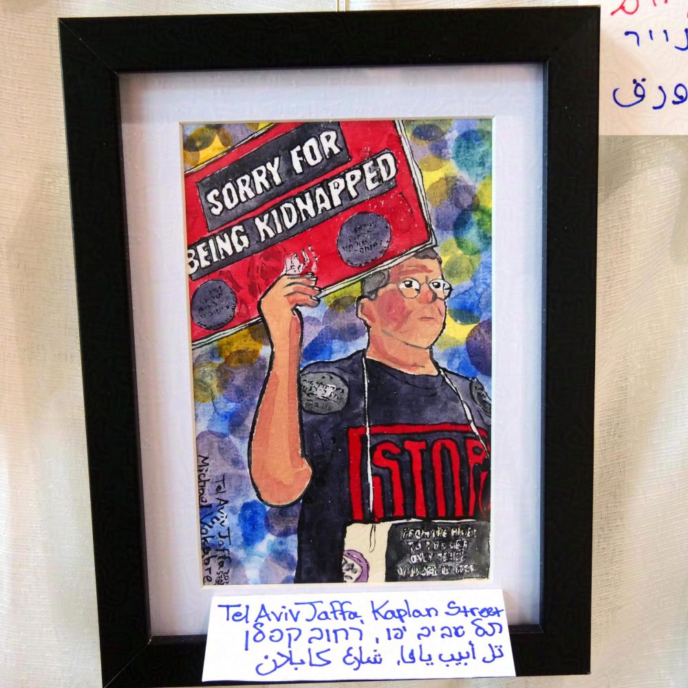 Vertical painting, a portrait of a man standing painted in half, looking to front-right, wearing glasses, holding a banner with his right hand on top of his head. The banner says "Sorry for being kidnapped" in white letters on black on red background. The man's shirt says "STOP" which morphs into "stop war", on the shoulders are two black stickers, and in the front, hanging by the neck is another banner with two purple stickers, and the text in the middle saying "from the river to the sea only peace will set us free". The background is painted in multiple circles of different colours. The painting is in black frame on white background, behind the frame is white cloth.