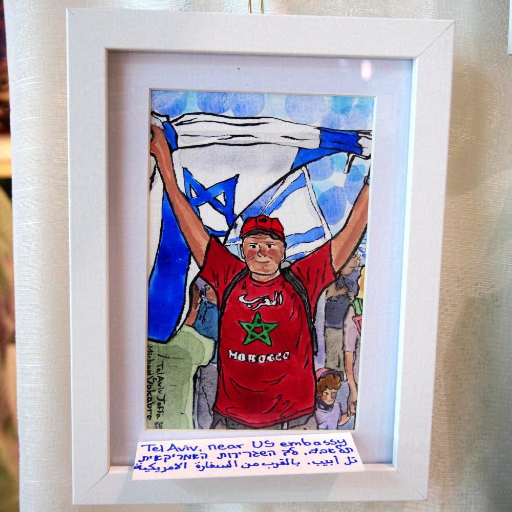 Vertical painting in watercolour with ink lines. In the middle is a middle-aged man in a t-shirt which has "Morocco" written in Arabic and English and Moroccan star in the middle, above his head he holds an Israeli flag. Behind are multiple more Israeli flags held by the crowd. On the right is a woman holding a kid by the hand. The painting is in white frame on white background, affixed to white cloth.