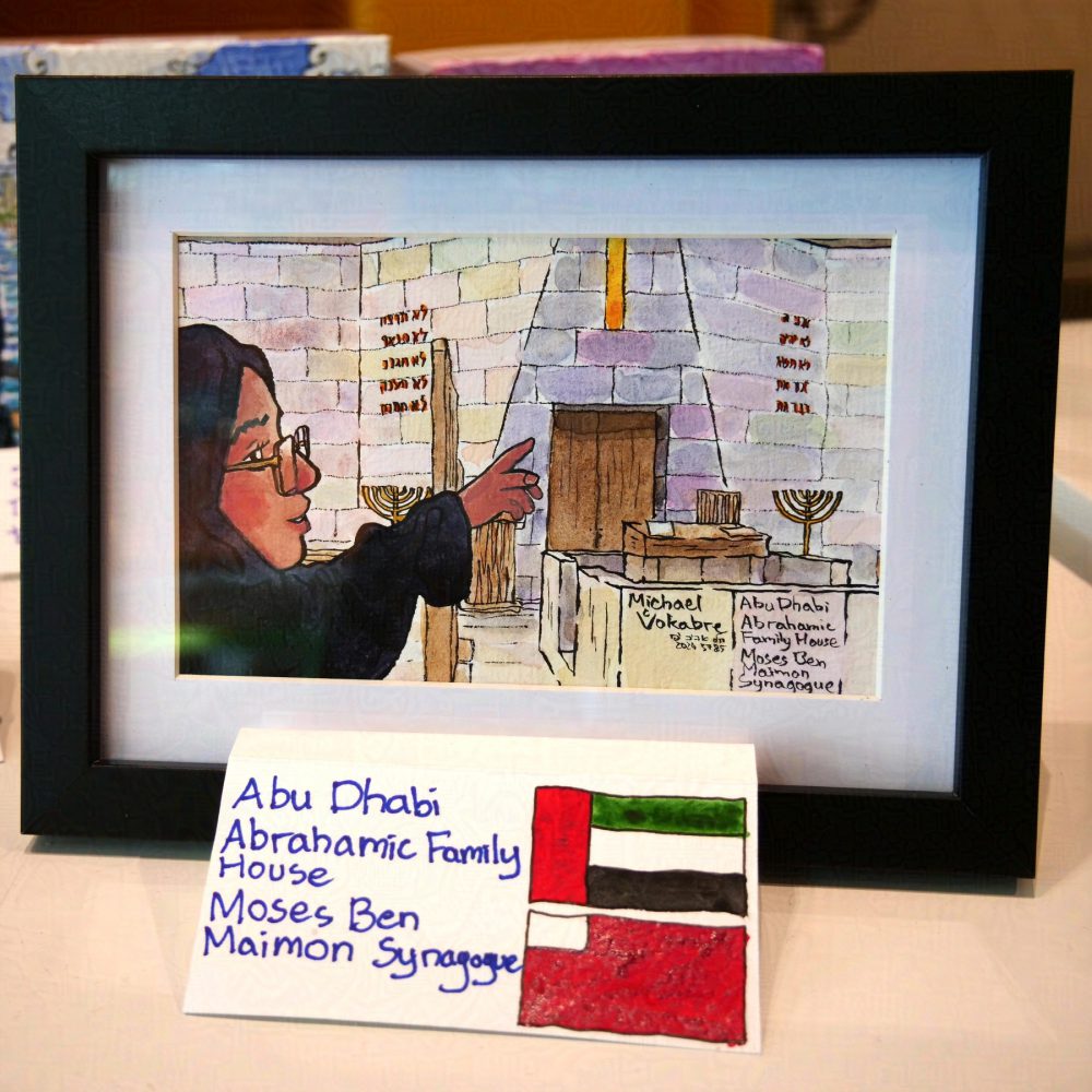 Horizontal aligned painting painted in watercolour in ink lines, showing the interior with the center of Moses Ben Maimon Synagogue at the Abrahamic Family House in UAE. On the left stands a hijabi guide, smiling, showing the place. On the right is bima, with a division fence on the left, behind one can see two hanukkiahs, and in the middle is the Torah niche. Above is the hole with a light. The walls are from white-ish stone tiles, painted in different colours, and above peeking are the windows. On the left and right from the center are ten commandments written in Hebrew.
The painting is on the white background in a black frame, below it's signed in English, with flags of UAE and Dubai to the right.