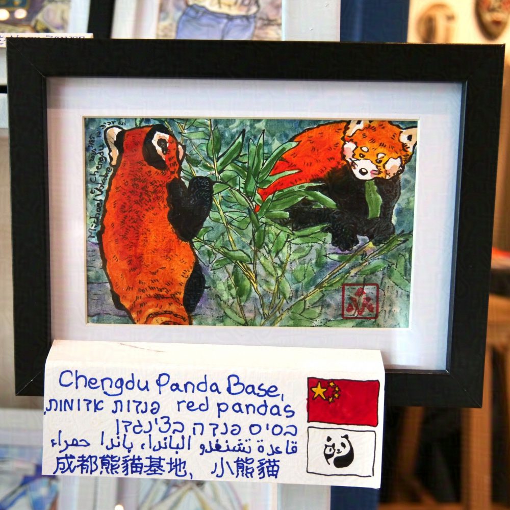 Horizontal painting, in ink with watercolour lines. On the left is a red panda holding a bamboo branch, to the back to the observer. On the right is another red panda on a stump chewing a bamboo leaf, part of the red panda obscured by the branch. Below is a plaque, hand-written, in English, Hebrew, Arabic and Chinese, with a flag of P.R.China and Chengdu Panda Base to the right. The painting is in a black frame with white background between the painting and the frame.  Behind are blurry artworks displayed alongside this piece. 