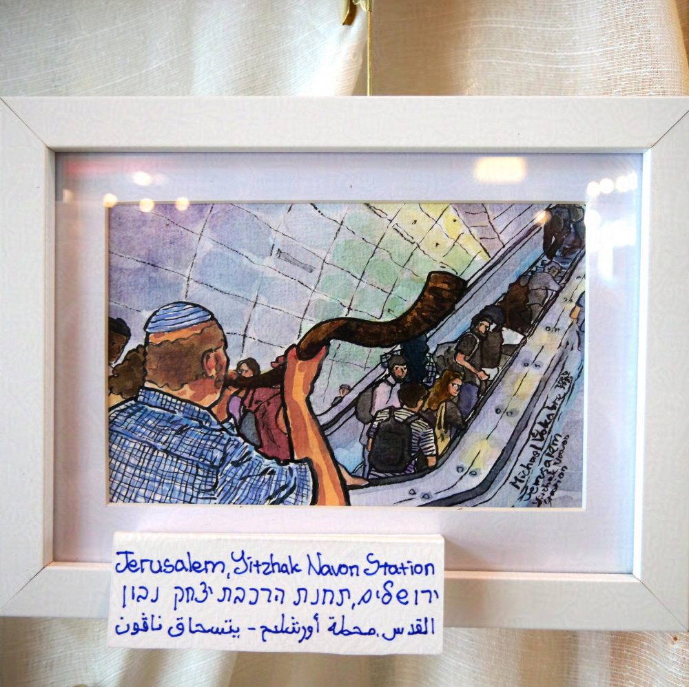 Horizontal aligned painting, in which from the left to the right there are two escalators, and in front on the left stands with his back to the observer, a bolding man in a kippah, blowing a shofar towards the escalators. Some people on the escalator look confused back. The painting is made in watercolours with ink lines, and the tiles of the wall are painted in multiple colours. The painting is in a white frame, on a white background, and has a signature below saying "Jerusalem, Yitzhak Navon Station" in English, Hebrew and Arabic.