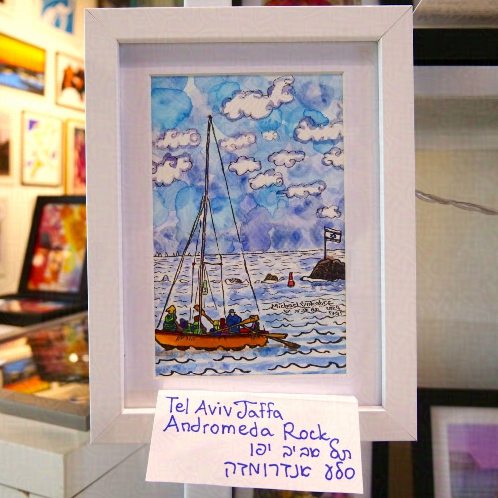 The painting described before is photographed framed affixed to the corner of a stall being sold at the port of Jaffa. In the background are other paintings. The frame is white, and in between the frame and the painting is white background. Below the painting is a sheet of paper with hand-written description of the painting in English and Hebrew.